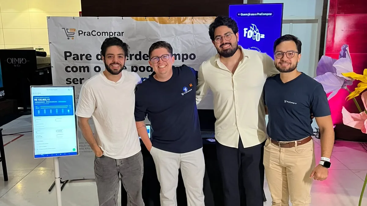 Equipe do PraComprar sorrindo em frente ao estande da empresa, com o banner da marca ao fundo e um totem exibindo o painel do sistema